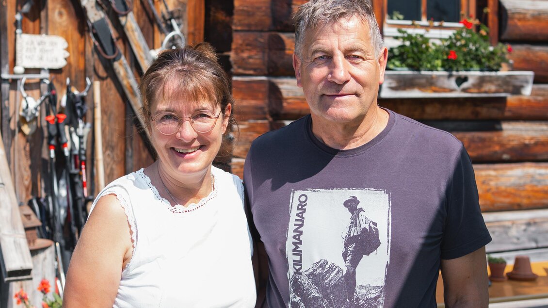 Ein freundliches Paar steht vor einer Holzfassade. Die Frau trägt ein weißes Oberteil und die Hose ist grün, während der Mann ein T-Shirt und eine grüne Schürze trägt. | © TV Gesäuse