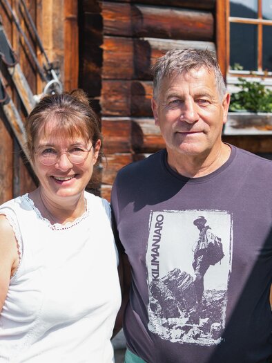 Ein freundliches Paar steht vor einer Holzfassade. Die Frau trägt ein weißes Oberteil und die Hose ist grün, während der Mann ein T-Shirt und eine grüne Schürze trägt. | © TV Gesäuse
