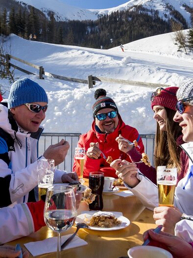 A group of friends is enjoying a meal outdoors in a snowy mountain landscape. They are wearing warm clothing and laughing together as they experience the winter atmosphere. | © Präbichl Bergbahnen