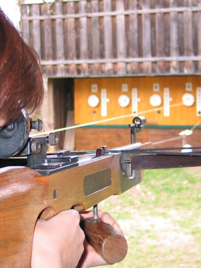 Eine Frau in traditioneller Kleidung steht vor einer Wand voller Teller und zielt mit einem historischen Gewehr. Sie lächelt und scheint Spaß daran zu haben, das Gewehr auszuprobieren. | © Armbrustschießen, Bad Aussee_Rastl