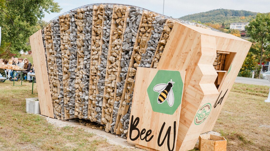 Ein innovatives Bienenhaus aus Holz und Steinen, das auf einem grünen Rasen steht. Es hat ein modernes Design mit einem Bienen-Symbol und einem einladenden Aussehen. | © MotionAds