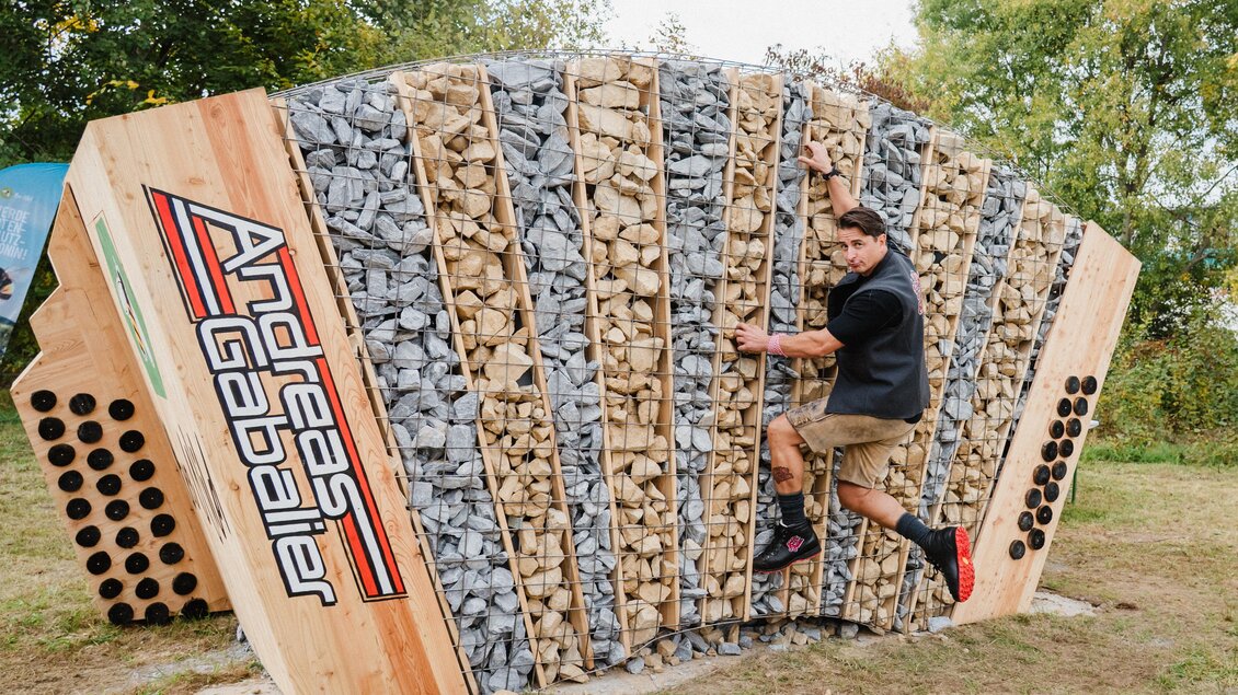 Ein Kletterer erreicht eine Wand aus Holz und Steinen. Die Wand ist bunt gestaltet und erinnert an ein Design von Andreas Gabalier. | © Bee Wild
