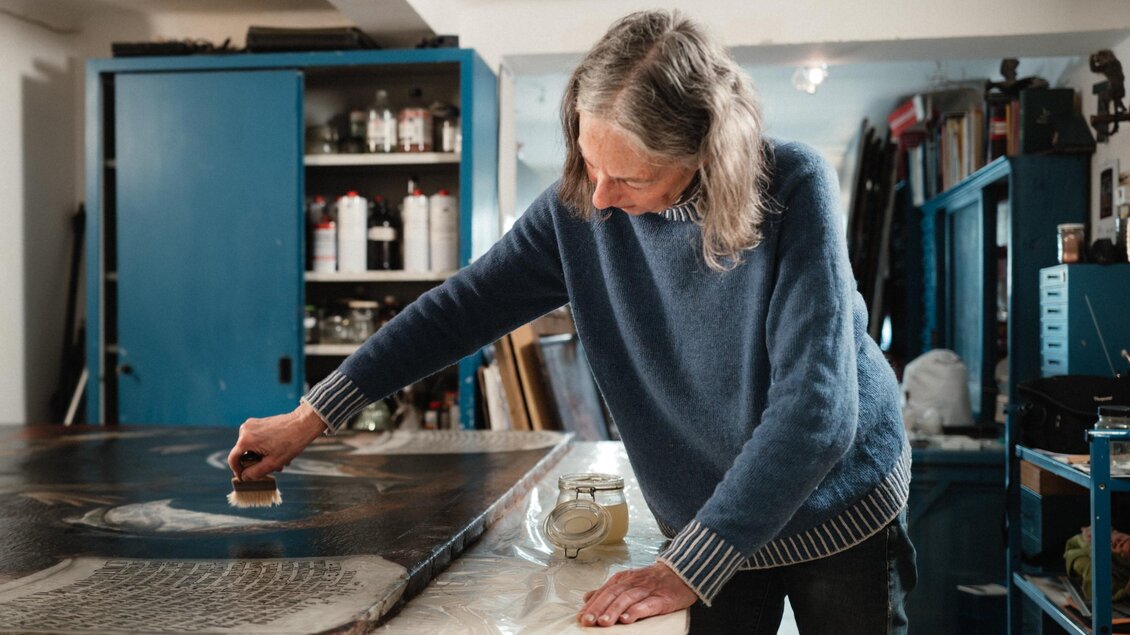 Restauratorin Erika Thümmel bearbeitet mit einem breiten Pinsel ein großformatiges Gemälde auf einem Arbeitstisch. | © Bernd Grosseck