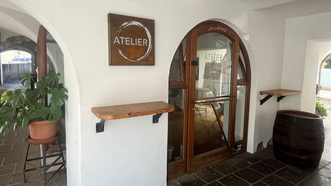 Ein einladender Eingangsbereich eines Ateliers mit einem Holzschild und großen Fenstern. Links steht eine Pflanze und rechts ein Fass. | © TVB Ausseerland Salzkammergut_Nathalie Schröttenhamer