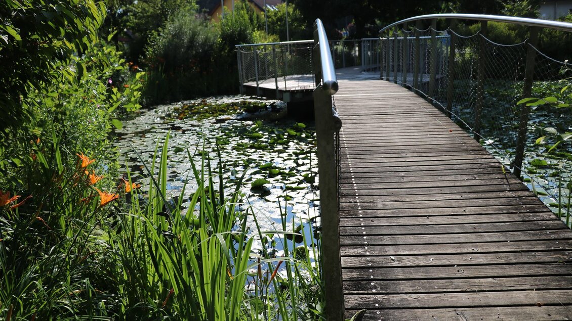 Eine Holzbrücke führt über einen Teich mit Seerosen. Umgeben von üppigem Grün und bunten Blumen ist die Szene friedlich und einladend. | © Julia Kerschbaumer