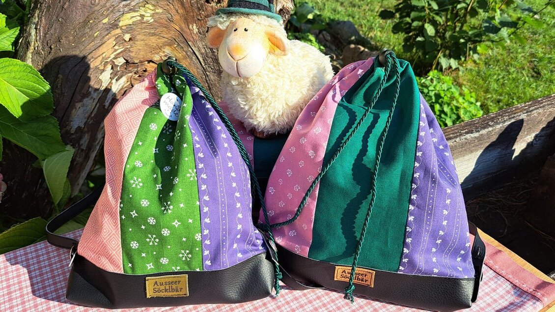 Zwei farbenfrohe Taschen stehen neben einem kuscheligen Schaf auf einem Tisch. Die Taschen sind mit verschiedenen Mustern in Grün, Pink und Lila gestaltet. | © Ausseer Söcklbär_Barbara Prentner
