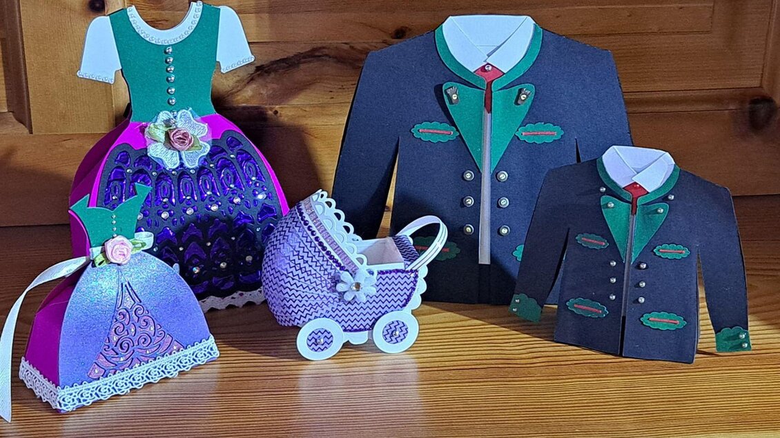 Eine Gruppe von dekorativen Kleidungsstücken aus Papier, inklusive einer Trachtenkleidung und einem Kinderwagen. Die Farben sind lebhaft, mit lila, grün und blau, und die Details sind kunstvoll gestaltet. | © Ausseer Söcklbär_Barbara Prentner