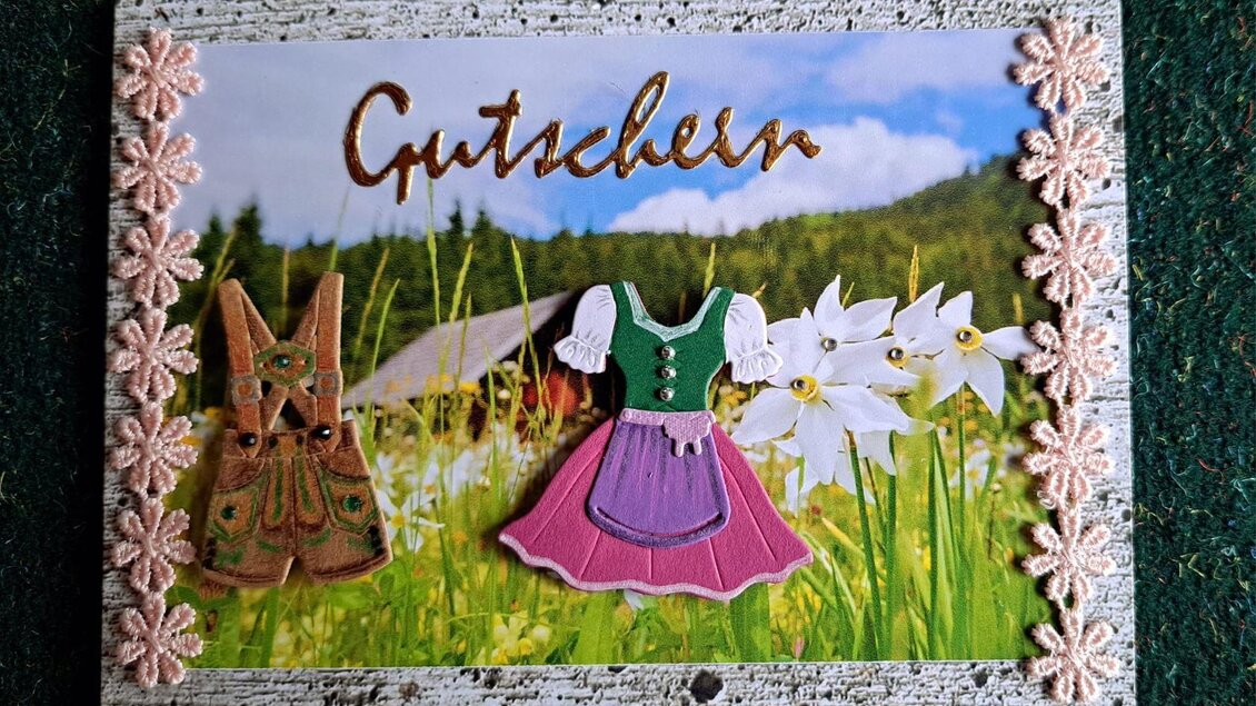 Eine farbenfrohe Karte mit dem Wort "Gutscheine" und Illustrationen eines Dirndls und einer Lederhose. Im Hintergrund sind Wiesen und Blumen zu sehen. | © Ausseer Söcklbär_Barbara Prentner