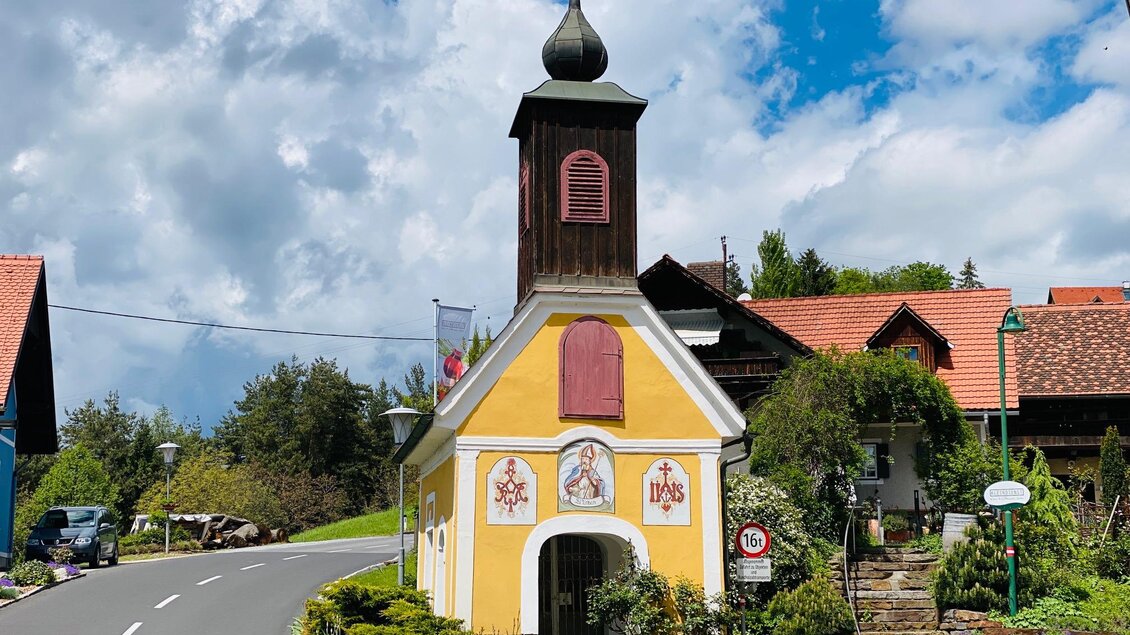 Eine bunte kleine Kapelle mit einem spitzen Turm steht an einer Straße. Im Hintergrund sind grüne Hügel und ein blauer Himmel mit Wolken zu sehen. | © TV Südsteiermark