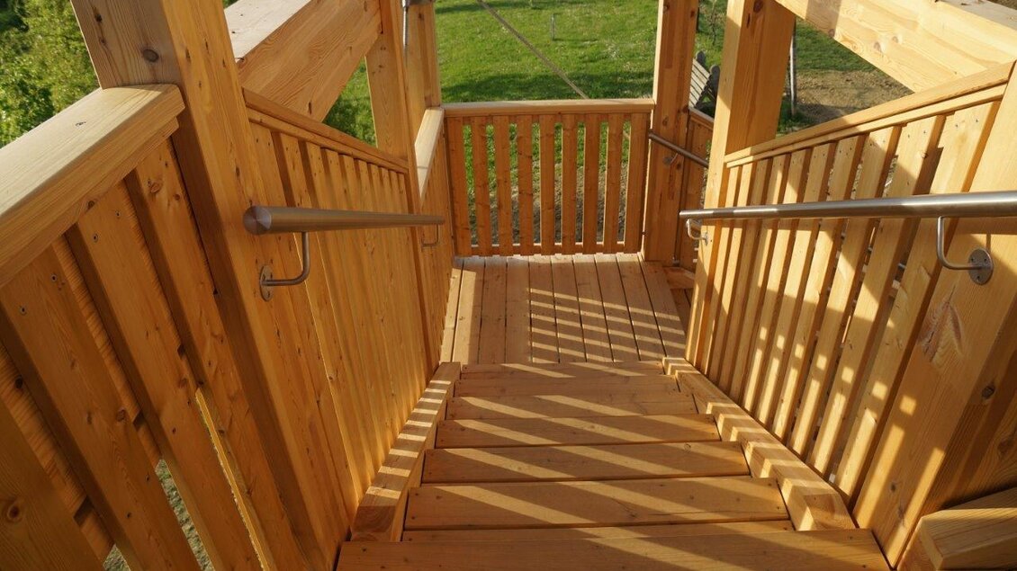 Eine hölzerne Treppe mit Geländer, die nach unten führt. Die Umgebung ist grün und es ist ein sonniger Tag. | © Gemeinde Unterlamm