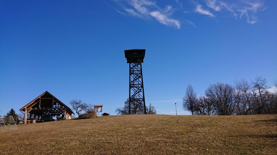 Ein Aussichtsturm auf einem sanften Hügel unter einem klaren blauen Himmel. Im Vordergrund ist eine kleine Hütte sichtbar. | © TV Südsteiermark