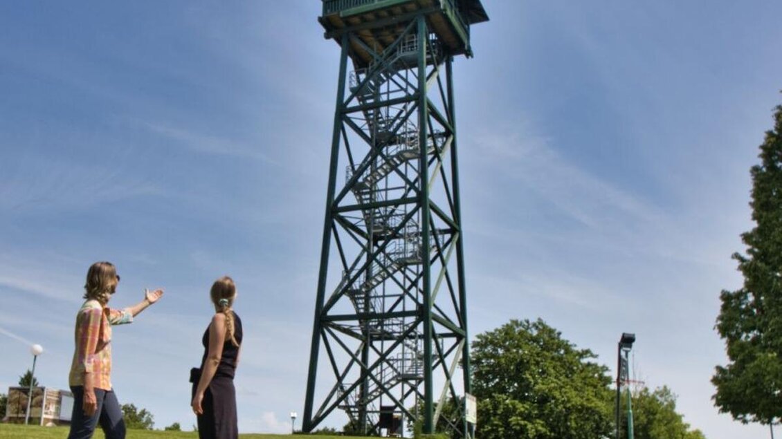 Zwei Frauen stehen auf einer Wiese und schauen auf einen Aussichtsturm. Der Himmel ist klar und es gibt viel grünes Gras und Bäume in der Umgebung. | © WEGES