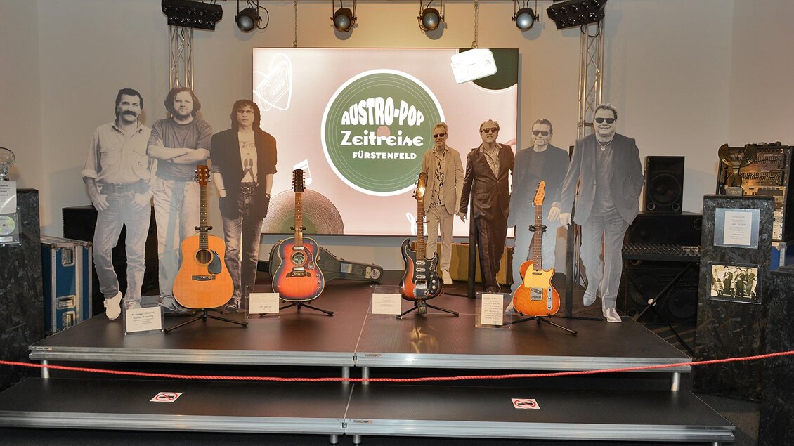 Eine Ausstellung mit Gitarren und lebensgroßen Figuren aus Papier. Im Hintergrund ist ein großes Bildschirm mit dem Schriftzug "Austro-Pop Zeitreise". | © Austro-Pop-Zeitreise
