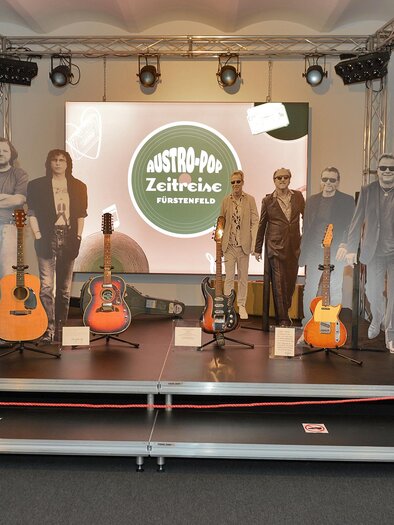 An exhibition with guitars and life-sized figures made of paper. In the background, there is a large screen with the inscription "Austro-Pop Time Travel". | © Austro-Pop-Zeitreise