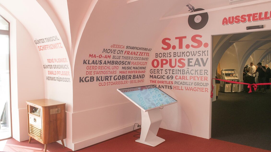 Ein heller Ausstellungsraum mit weißen Wänden und roten Akzenten. An der Wand stehen Namen von Künstlern und Bands, während ein interaktives Display im Vordergrund zu sehen ist. | © Austro-Pop-Zeitreise