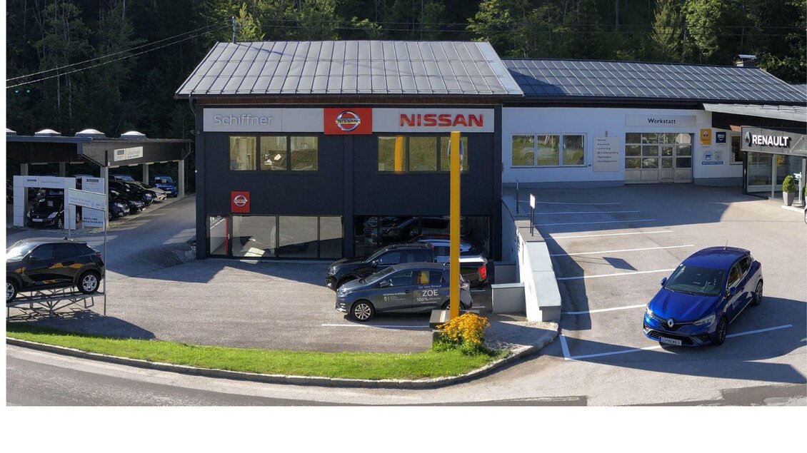 Ein modernes Autohaus mit mehreren PKW auf dem Parkplatz. Die Gebäude sind freundlich und gut beleuchtet, umgeben von Bäumen. | © Autohaus Schiffner