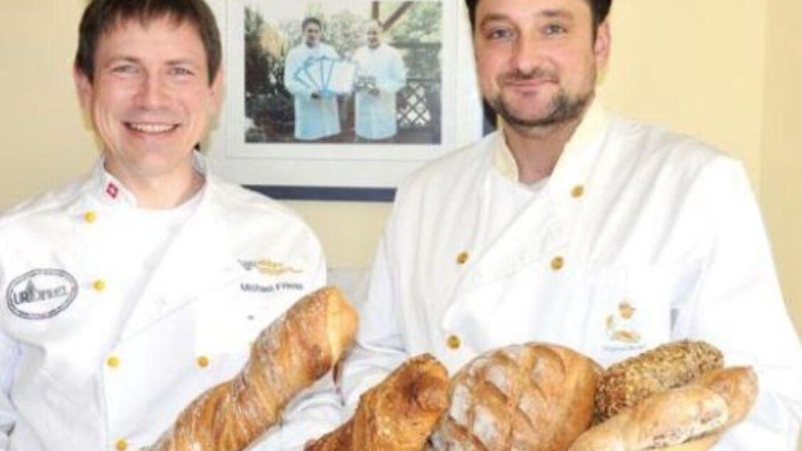 Zwei Bäcker in weißen Kitteln halten frisches Brot in den Händen. Im Hintergrund ist ein Bild von ihnen zu sehen. | © Bäckerei Altenburger