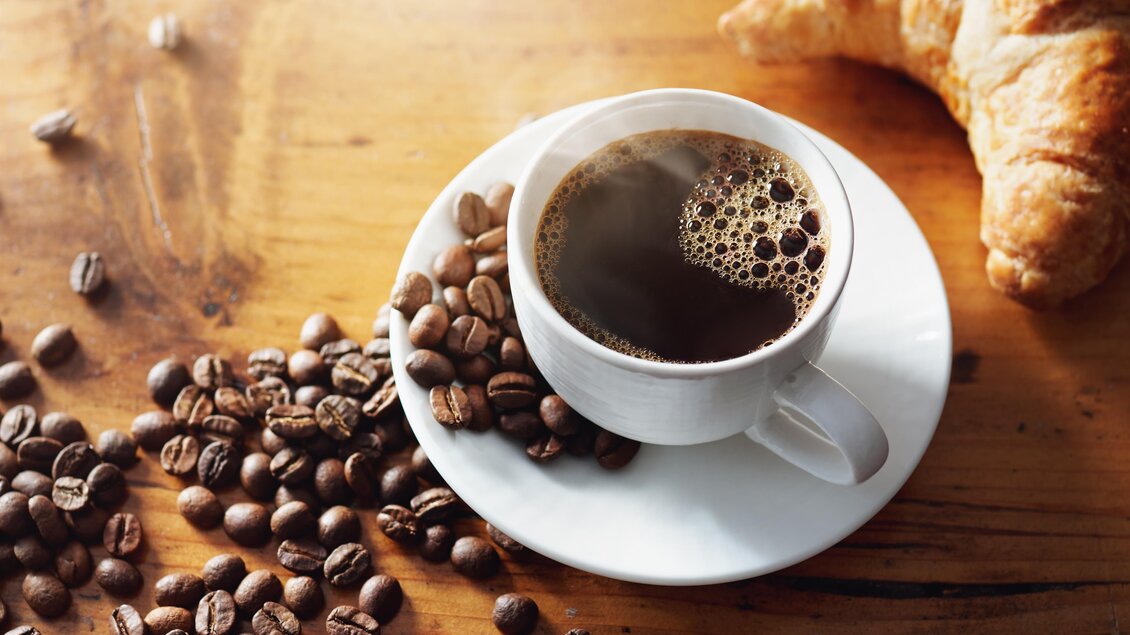 Eine Tasse Kaffee auf einem Unterteller, umgeben von Kaffeebohnen. Neben der Tasse liegt ein Croissant. | © AdobeStock_270222935_Cafehaus