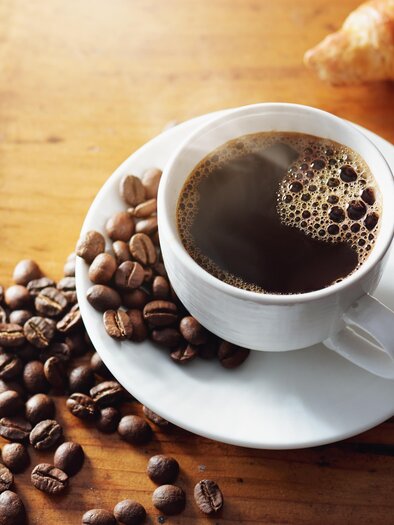 Eine Tasse Kaffee auf einem Unterteller, umgeben von Kaffeebohnen. Neben der Tasse liegt ein Croissant. | © AdobeStock_270222935_Cafehaus