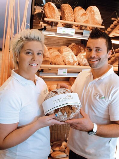 Ein freundliches Team präsentiert frisches Brot in einer Bäckerei. Im Hintergrund sind verschiedene Brotsorten zu sehen. | © Bäckerei Masser