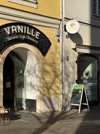 An inviting café named "Vanille" with a stylish entrance.  
In front of the café, there is a cozy chair and a board with offers. | © TV Hochsteiermark