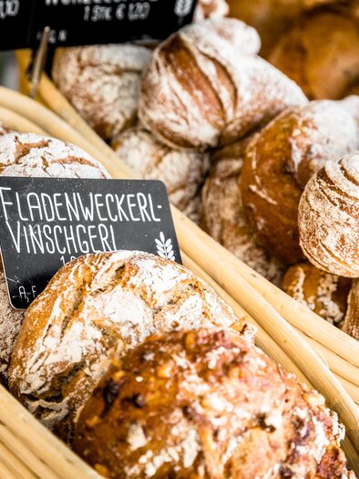 Ein Korb mit frisch gebackenem Brot und Brötchen, darunter Fladenweckerl. Die Backwaren sind mit Puderzucker bestäubt und in einer Bäckerei präsentiert. | © Stephan Friesinger