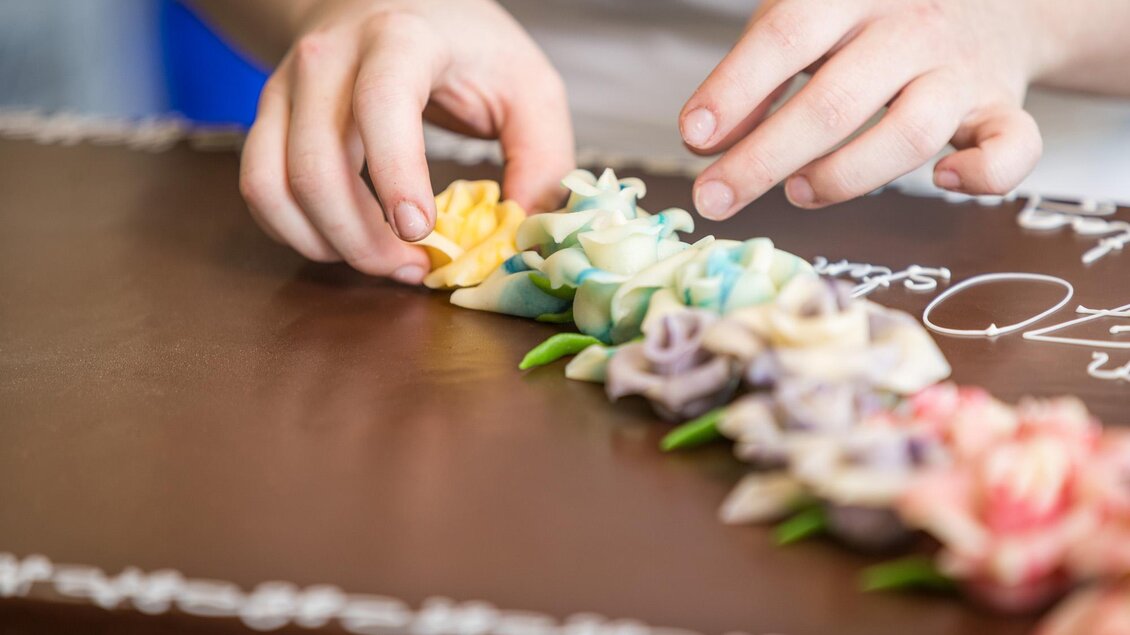 Eine Person dekoriert einen Schokoladenkuchen mit bunten Rosen aus Zuckerguss. Die detailreiche Verzierung zeigt verschiedene Farben und Formen. | © Netzwerk Kulinarik wildbild