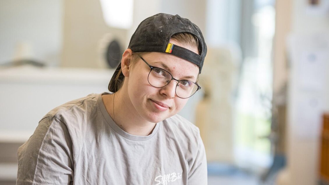 Eine Person arbeitet an einer dekorativen Torte in einem hellen Raum. Im Hintergrund sind weitere Gegenstände und eine Uhr sichtbar.