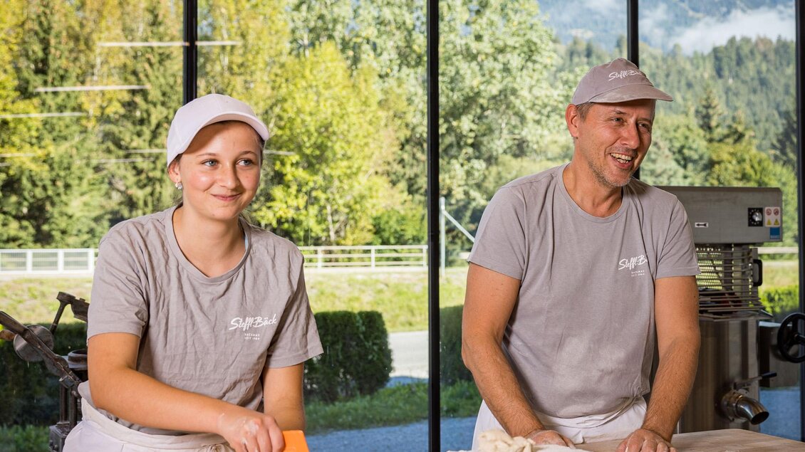 Zwei Bäcker arbeiten in einer modernen Bäckerei. Im Hintergrund sind Berge und ein grüner Wald zu sehen. | © Netzwerk Kulinarik wildbild