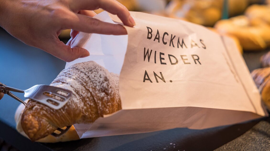Ein Croissant wird aus einer Papiertüte genommen, die mit "BACKMAS WIEDER AN." beschriftet ist. Im Hintergrund sind weitere Backwaren sichtbar. | © Netzwerk Kulinarik wildbild
