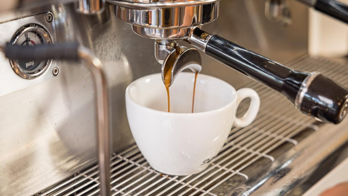 Ein Kaffee wird frisch aus einer Espressomaschine in eine weiße Tasse gegossen. Die Maschine hat ein glänzendes Edelstahl-Finish. | © Netzwerk Kulinarik wildbild
