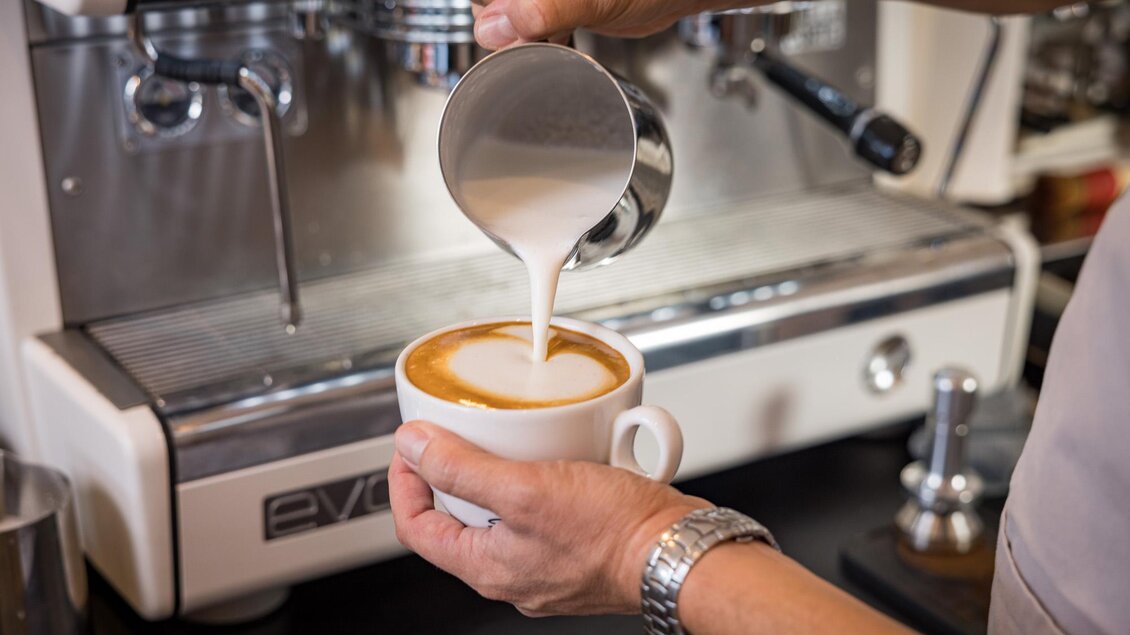 Ein Barista gießt Milch in eine Tasse Kaffee. Im Hintergrund ist eine Espressomaschine zu sehen. | © Netzwerk Kulinarik wildbild