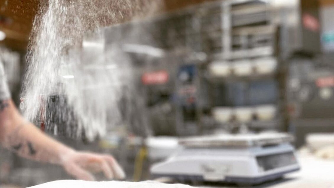 Ein Bäcker streut Mehl über einen Teig auf einem Tisch. Im Hintergrund sind weitere Küchengeräte sichtbar. | © Bäckerei Steffl-Bäck