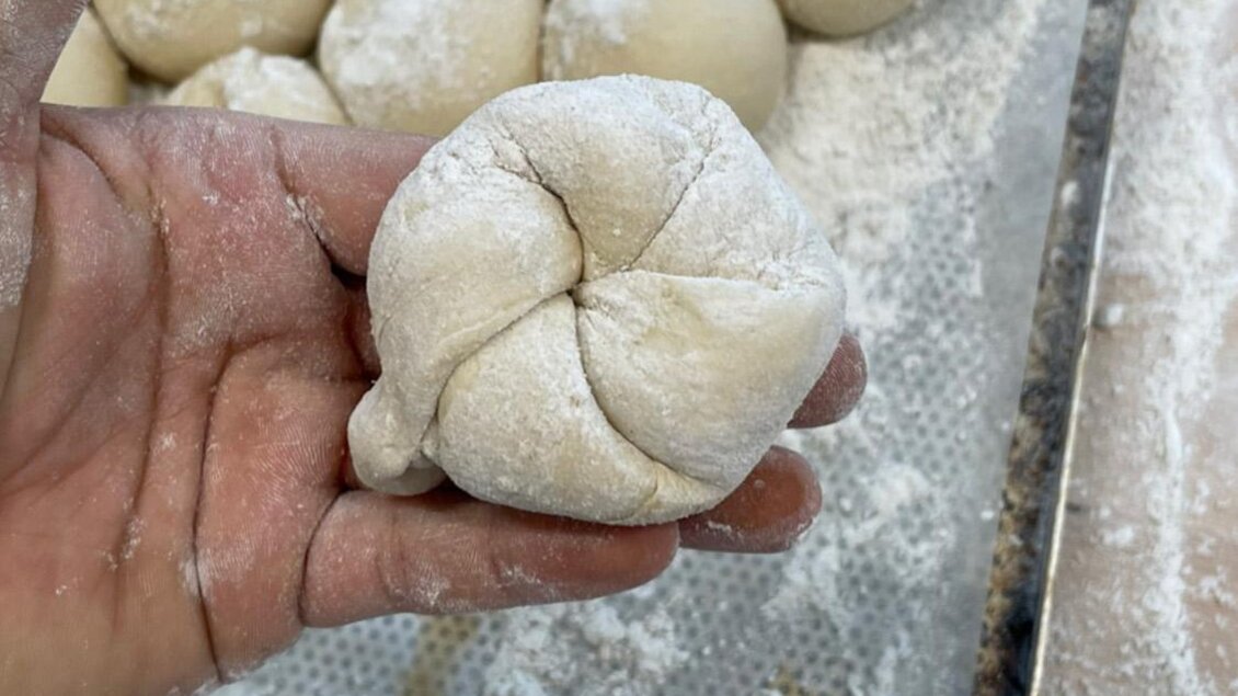 Eine Hand hält einen geformten Teigball. Im Hintergrund sind weitere Teigbälle auf einer bemehlten Fläche zu sehen. | © Bäckerei Steffl-Bäck
