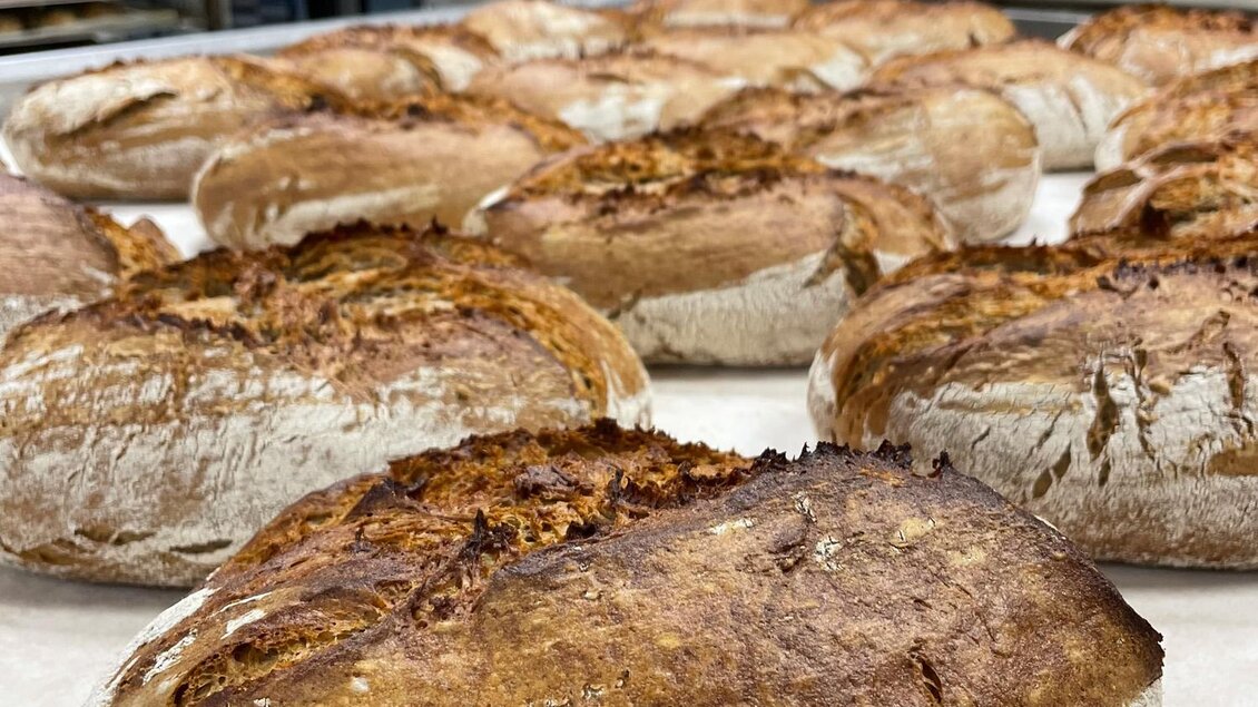 Frische, goldbraun gebackene Brote liegen auf einem Blech in einer Bäckerei. Im Hintergrund ist ein Bäcker zu sehen, der weitere Brote aus dem Ofen holt. | © Bäckerei Steffl-Bäck
