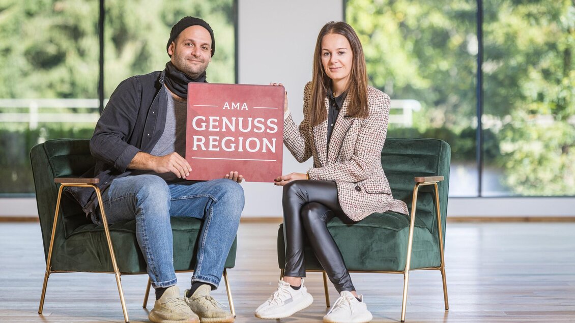 Zwei Personen sitzen auf Sesseln und halten ein Schild mit der Aufschrift "AMA GENUSS REGION". Der Hintergrund zeigt große Fenster mit viel Tageslicht. | © Netzwerk Kulinarik wildbild