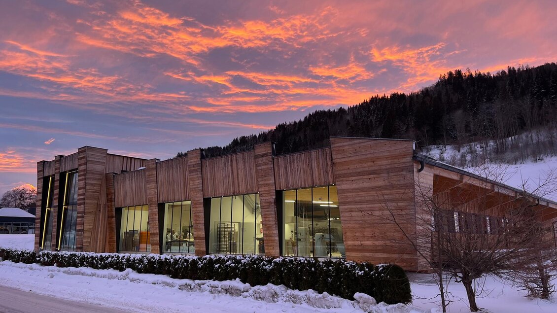 Ein modernes Gebäude aus Holz vor einer verschneiten Landschaft. Der Himmel ist in warmen Farben während des Sonnenuntergangs beleuchtet. | © Bäckerei Steffl-Bäck