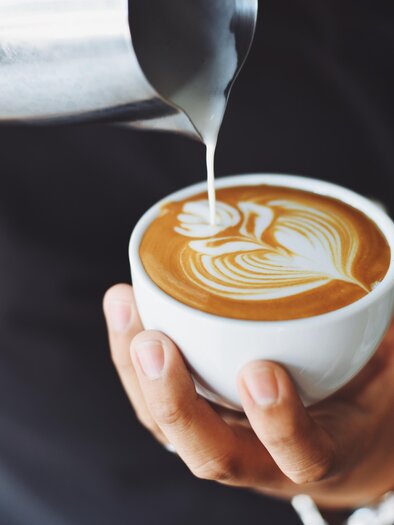 Eine Person gießt Milch in eine Tasse mit Kaffee. Auf der Oberfläche ist ein kunstvolles Muster zu sehen. | © AdobeStock_131053005_Cafehaus