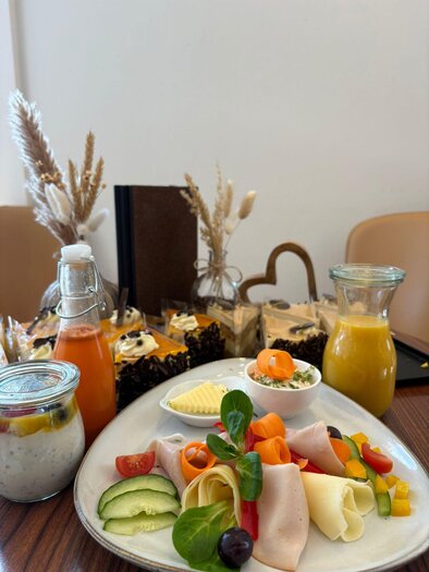 Ein reichhaltiger Frühstückstisch mit frischem Obst, Gemüse und Wurstaufschnitt. Im Hintergrund sind verschiedene Gebäcke und Getränke zu sehen.