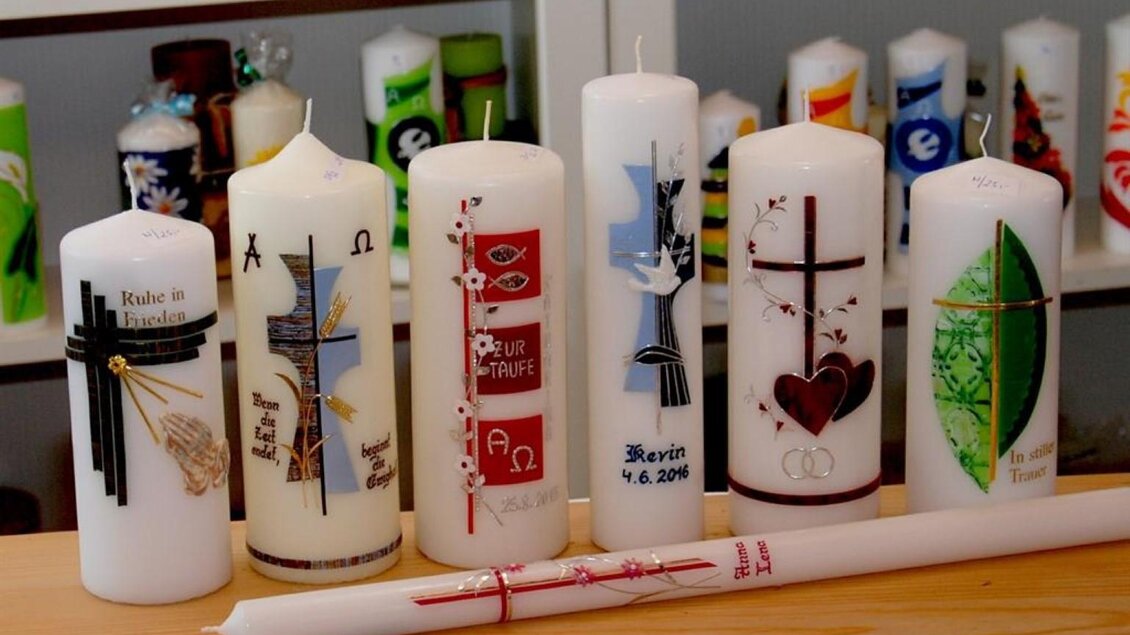 Eine Reihe von dekorativen weißen Kerzen mit verschiedenen religiösen und festlichen Motiven. Die Kerzen sind auf einem Holzregal angeordnet und zeigen bunte Verzierungen. | © Bäuerliches Klein- und Kunsthandwerk