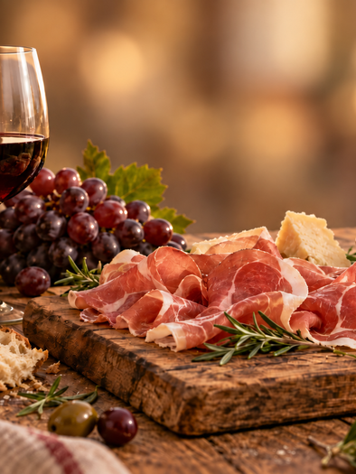 Board with ham, bread, grapes, and olives next to a glass of red wine | © ChatGPT Image_Astrid Lackner