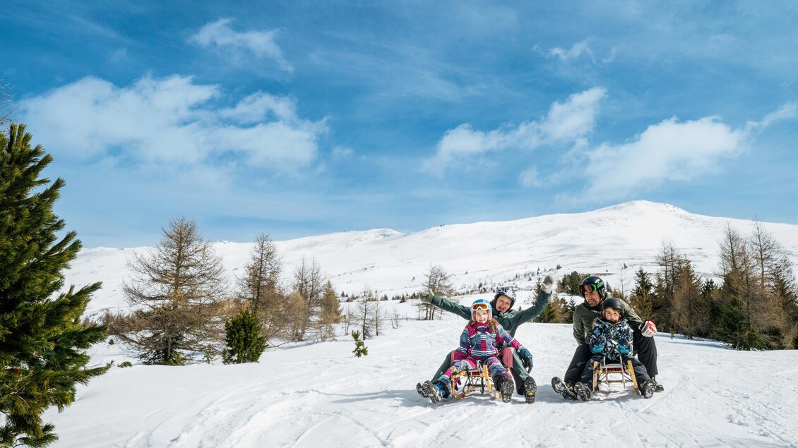 Eine Gruppe von Menschen fährt auf Schlitten im verschneiten Gelände. Im Hintergrund sind schneebedeckte Berge und ein blauer Himmel zu sehen. | © TV Murau