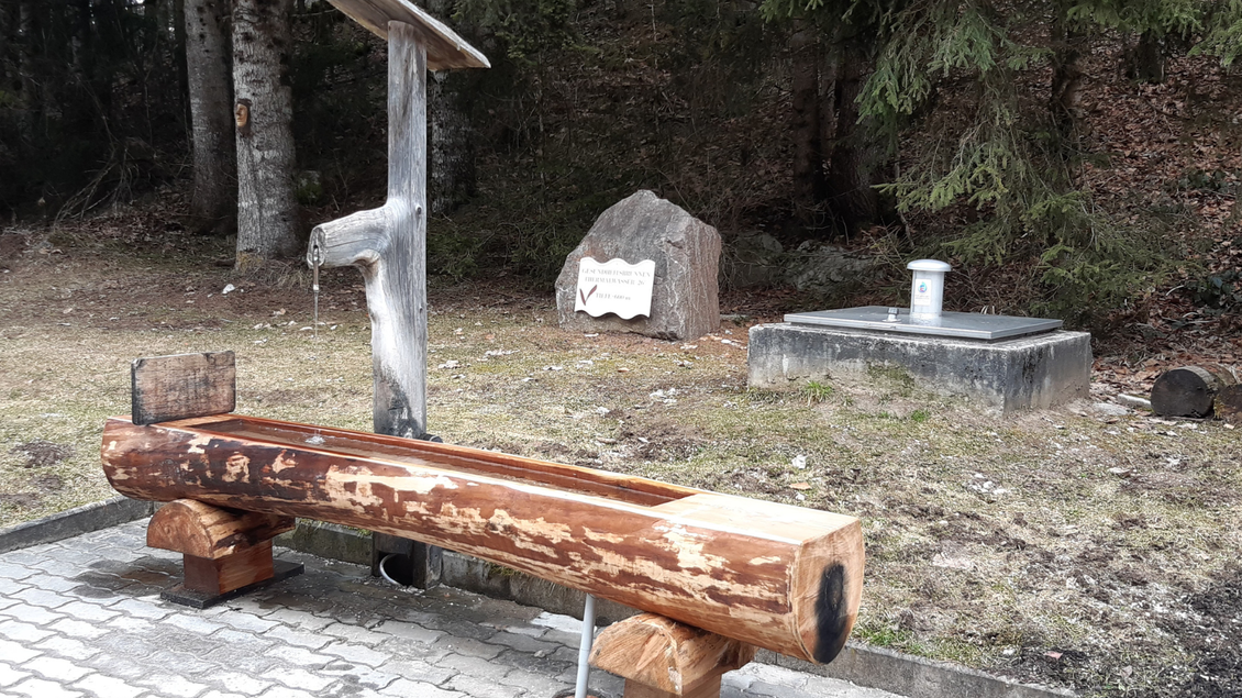 Eine Holzbank und ein Holzbrunnen in einem ruhigen Waldgebiet. Im Hintergrund sind Bäume und ein Felsen mit einer Beschriftung zu sehen. | © TVB Ausseerland Salzkammergut_Daniela Casari