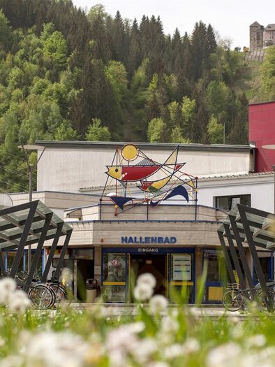 Ein modernes Gebäude mit einem bunten Kunstwerk an der Fassade. Im Vordergrund wachsen Löwenzähne auf einer grünen Wiese.