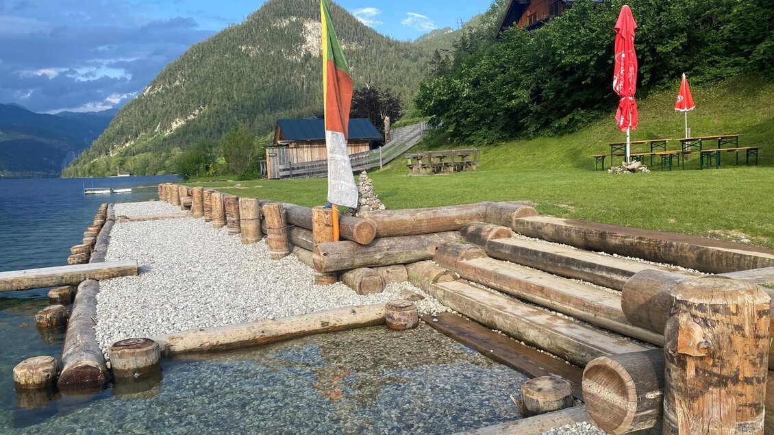 <p>Der Weg aus Holz mit Kies führt ins klare Wasser des Grundlsees, umgeben von einer grünen Wiese. Im Hintergrund sind Tische mit Bänken und Schirmen zu sehen.</p> | © Bettina Scheck
