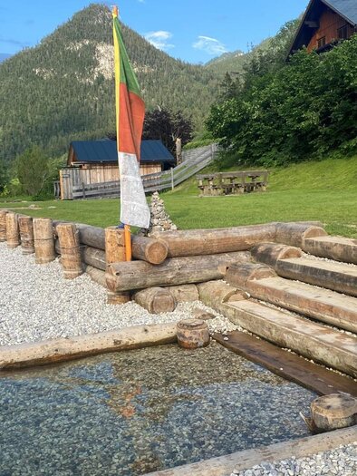 <p>The wooden path with gravel leads to the clear water of Grundlsee, surrounded by a green meadow. In the background, tables with benches and umbrellas can be seen.</p> | © Bettina Scheck