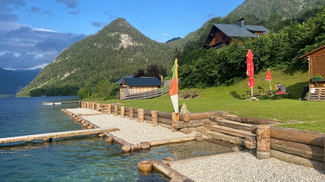 <p>Der Grundlsee mit dem Seezugang aus Holz und Kies und einem Blick bis nach Gößl. Im Hintergrund sind gemütliche Sitzmöglichkeiten und Sonnenschirme zu sehen.</p> | © Bettina Scheck