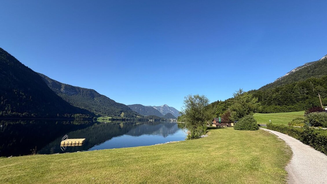 <p>Der ruhige Grundlsee umgeben von den majestätischen Bergen und Wiesen. Der Himmel ist blau und klar. Auch die Liegewiese vom Badeplatz Gaiswinkel ist bietet ausreichend Platz für zahlreiche Badegäste.</p> | © Tourismusverband Ausseerland
