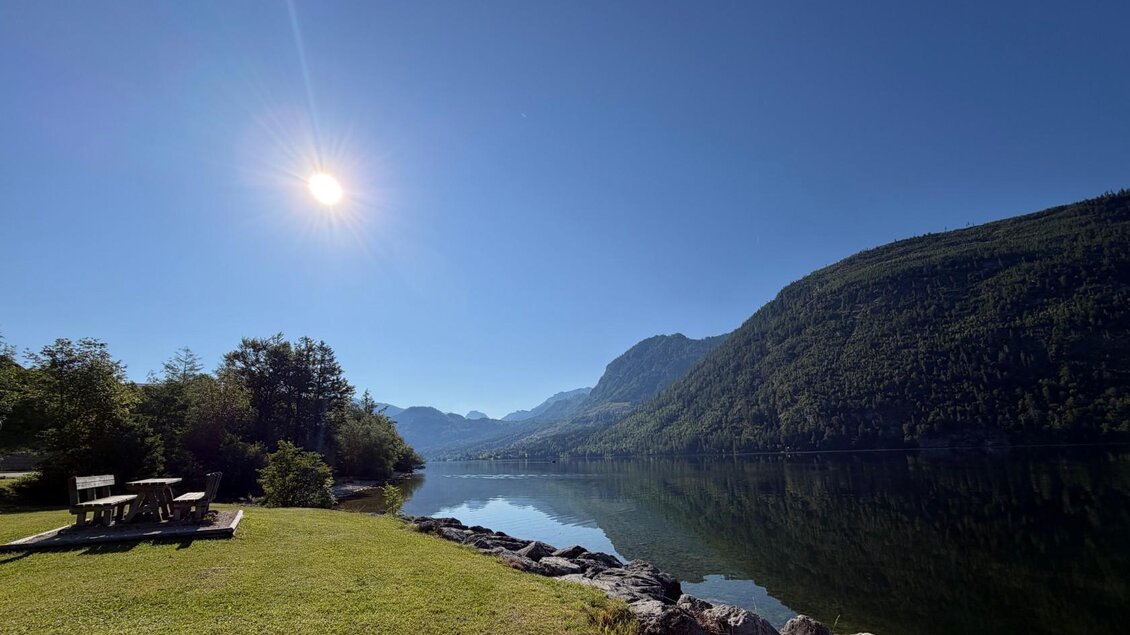 Die idyllische Landschaft am Grundlsee mit der Sonne über dem Toten Gebirge. Im Vordergrund steht eine gemütliche Bank auf einer saftig grünen Wiese. | © Tourismusverband Ausseerland