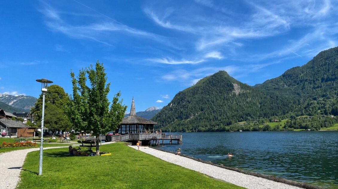 <p>Der glasklare Grundlsee umgeben vom majestätischen Toten Gebirge und einer saftigen Grünfläche. Im Vordergrund ist der Weg durch den Park und der Musikpavillon zu sehen.</p> | © Tourismusverband Ausseerland